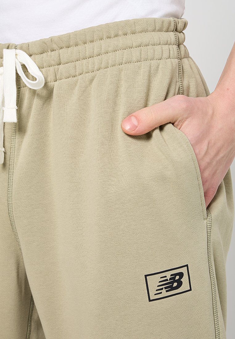 Pantalons de survêtement beiges avec taille élastique et cordon de serrage. Dispose de poches latérales et d'un patch logo "NB" noir sur la cuisse gauche. Texture douce.
