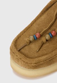 Chaussure à enfiler en daim marron, ornée de perles décoratives rouges, bleues et beige sur un lacet tressé, avec une semelle texturée et une forme arrondie.