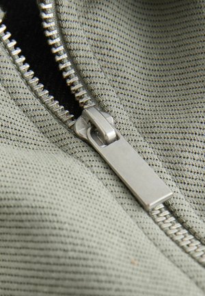 Dragkedjedetalj på texturerat, dämpat grönt tyg; metallskjutregel med flat yta och synliga tänder längs ena sidan, som visar funktionell design.