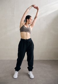 Barna sportmelltartó logóval, fekete, rugalmas szegélyű melegítőnadrág és fehér sportcipő. A téma nyújtózkodik egy semleges háttér előtt.
