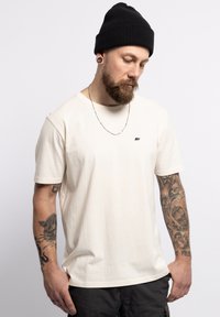 T-shirt di cotone beige con maniche corte, con un piccolo logo di un orso nero sul petto. Accompagnato da un berretto di maglia nero e una collana a catena.