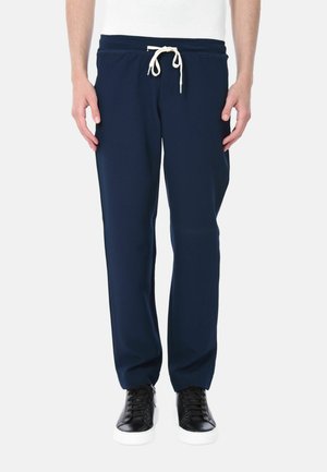 Uomo con pantaloni blu navy con coulisse e scarpe da ginnastica nere con suola bianca, in piedi davanti a sfondo bianco uniforme.