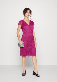 Vestido de renda fúcsia com mangas curtas em formato de onda, padrões florais e uma cintura ajustada. Combinado com sandálias de salto bloco pretas e uma clutch prateada.