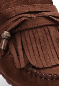 Mocasin de ante marrón con detalles de flecos, que presenta un acento de cuero trenzado y una textura de superficie suave. Vista en primer plano de los elementos de diseño.