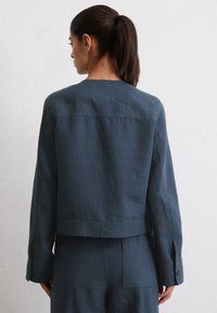 Donkerblauwe cropped jas van gestructureerde stof, met een eenvoudig ontwerp, lange mouwen en knoopsluitingen op de manchet. De achterkant benadrukt de vorm.