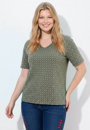 Lächelnde Frau mit langen blonden Haaren trägt grünes, gemustertes V-Ausschnitt-Shirt und blaue Jeans mit roter Blumenstickerei, steht vor weißem Hintergrund.