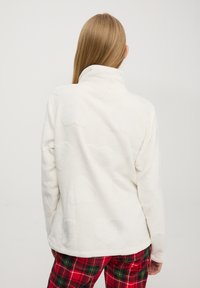 Pullover bianco in pile con collo alto, caratterizzato da morbidi motivi a cuore di diverse dimensioni. Abbinato a pantaloni a quadri rossi e verdi.