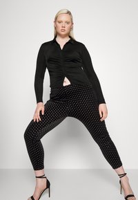 Camisa preta de mangas compridas com detalhe franzido, combinada com leggings pretas de veludo com padrão de bolinhas. O modelo usa saltos altos pretos.