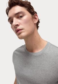 Šedé bavlněné tričko s kulatým výstřihem, krátkými rukávy a hladkým povrchem. Hlava modelu mírně nakloněná, neutrální pozadí.