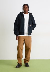Young man standing on green floor against white wall, wearing black hooded jacket, white t-shirt, brown pants, and black shoes.