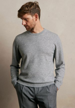 Szary sweter z dzianiny z długimi rękawami, okrągłym dekoltem i ściągaczami przy mankietach, zestawiony z jasnoszarymi spodniami. Miękka tekstura z delikatnymi wzorami.