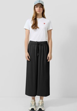 Młoda kobieta w białym t-shircie z małym czerwonym sercem, czarnych, szerokich spodniach, masywnych butach sportowych i jasnoniebieskiej dżinsowej czapce na jednolitym tle.