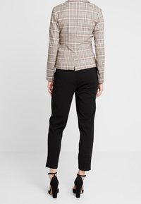 Blazer beige à carreaux avec une silhouette ajustée, associé à un pantalon noir sur mesure. Des chaussures noires à talons hauts complètent la tenue.