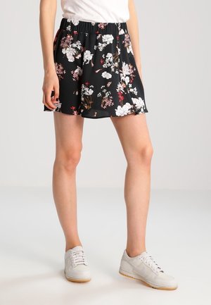Femme portant un short noir à fleurs, un t-shirt blanc et des baskets blanches, debout devant un fond gris clair uni.