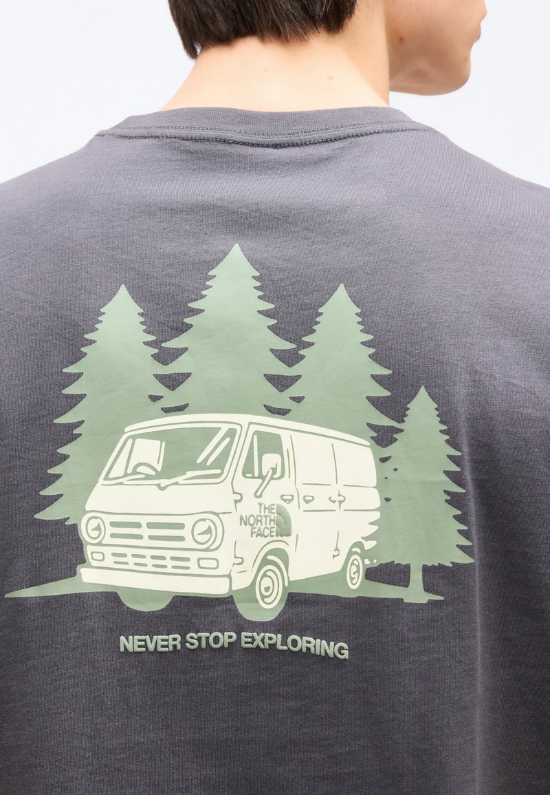 Person trägt ein graues Hemd mit einer Grafik eines Vans, Kiefern und dem Text "Never Stop Exploring" auf dem Rücken.