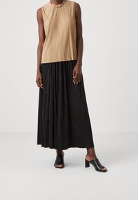 Blusa sin mangas en beige con pliegues verticales, combinada con una falda larga negra con pliegues. Mules de tacón abierto y negro.