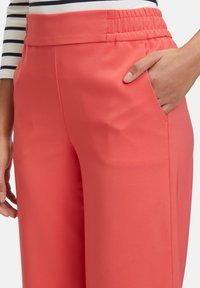 Coral, lightweight trousers with an elastic waistband, side pocket, and smooth texture. Visible stitching along the seams.