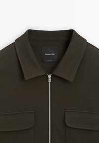 Mørkegrøn jakke med en struktureret krave, lynlåslukning og to brystlommer. Stoffet har et subtilt tekstureret mønster. Brandlabel synlig.