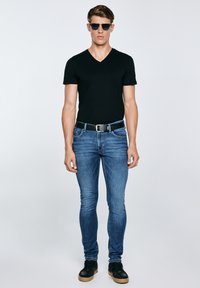 Fiatal férfi fekete V-nyakú pólóban, kék slim-fit farmerral, fekete övvel, fekete napszemüveggel és fekete cipőben, egyszínű háttér előtt állva.