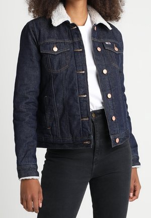 Vrouw draagt een donkerblauwe spijkerjas met witte fleece kraag, wit overhemd en zwarte broek, staand tegen een effen achtergrond.