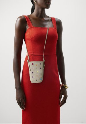 Bolso de ante beige con adornos coloridos, incluyendo estrellas y gemas, llevado con una correa al hombro. Modelo con un vestido rojo ajustado.