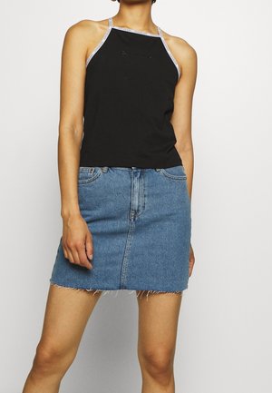 Kvinde iført sort haltertop med hvid kant og blå denimnederdel med frynset kant står mod en ensfarvet baggrund.