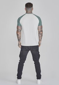 Tricou raglan alb și verde, realizat din țesătură, asociat cu pantaloni cargo negri, cu buzunare laterale. Persoana este văzută din spate.