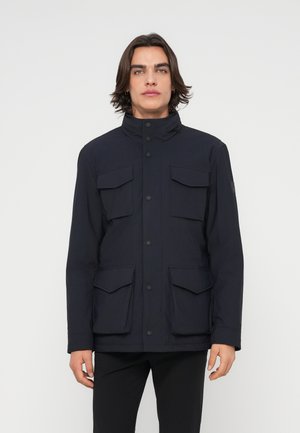 Homme aux cheveux mi-longs portant une veste noire avec quatre poches à rabat à l'avant et un col boutonné, debout devant un fond blanc.