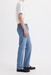 Jeans rectos de mezclilla de color azul claro, combinados con una camiseta ajustada blanca. Presenta una parche de cuero marrón en la parte trasera de la cintura.