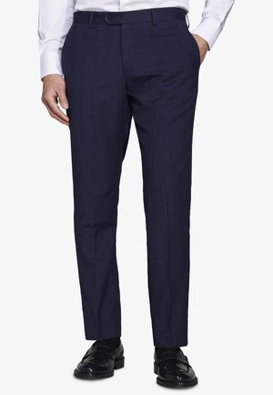 Uomo che indossa pantaloni da abito su misura blu navy scuro con motivo a quadretti discreto, abbinati a mocassini in pelle nera e camicia bianca.