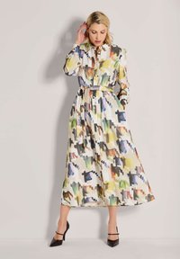 Eine Frau, die sich die Haare zurechtmacht und ein langes, knopfbestärktes Kleid mit mehrfarbigem abstraktem Muster und Gürtel trägt. Sie steht vor einem schlichten Hintergrund und trägt schwarze Absätze.