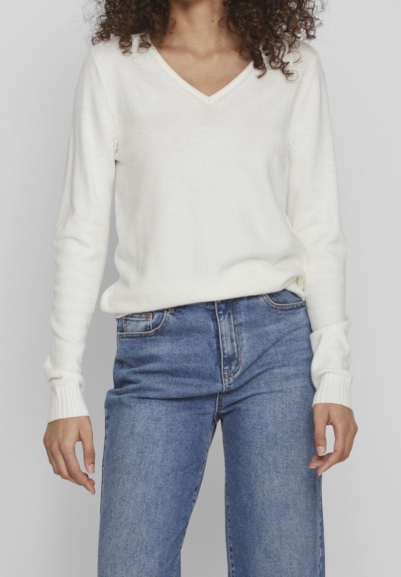 Witte, zachte gebreide trui met een V-hals, lange mouwen en ribgebreide manchetten. Gecombineerd met high-waisted blauwe jeans, wat een casual stijl uitstraalt.