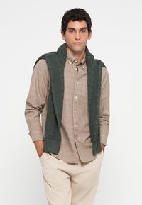 Man wearing beige checkered button-up shirt with dark green knit sweater draped over shoulders and light beige pants, hands in pockets.