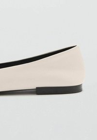 Zapato plano de cuero beige con suela negra baja, diseño de punta afilada y detalles de costura mínimos sobre un fondo gris claro.