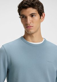 Light blue, ribbed sweater with a round neckline and embroidered logo on the chest. Soft texture and casual design suitable for layering.