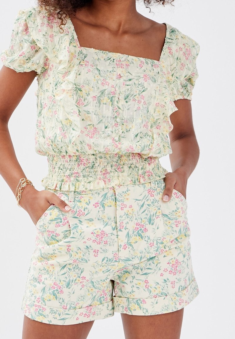 Femme portant un ensemble fleuri assorti avec un haut à manches courtes volantées et un short taille haute avec poches, mains dans les poches, bracelet en or au poignet gauche.