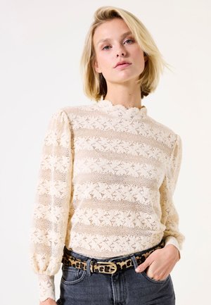 Blouse en dentelle crème, présentant des motifs rayés et un col en forme de coquille. Manches longues et bouffantes avec un tissu texturé et léger.