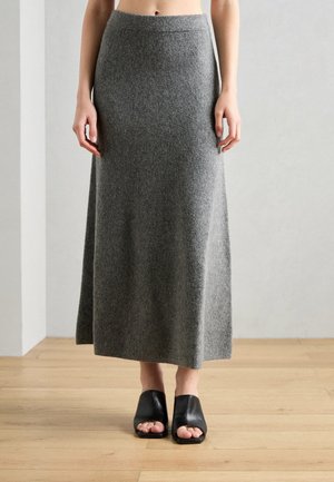 Kobieta ubrana w szarą, wełnianą spódnicę midi z wysokim stanem, zestawioną z czarnymi, otwartymi na palce butami na podwyższeniu, stojąca na jasnej, drewnianej podłodze.