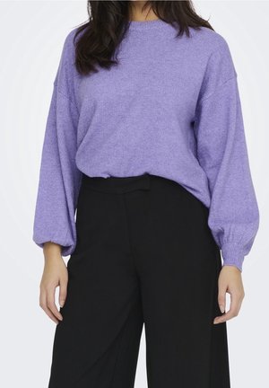 Vrouw draagt een losse lavendelkleurige trui met ballonmouwen, ingestopt in hoge zwarte wijde pijpenbroek, staand tegen een witte achtergrond.