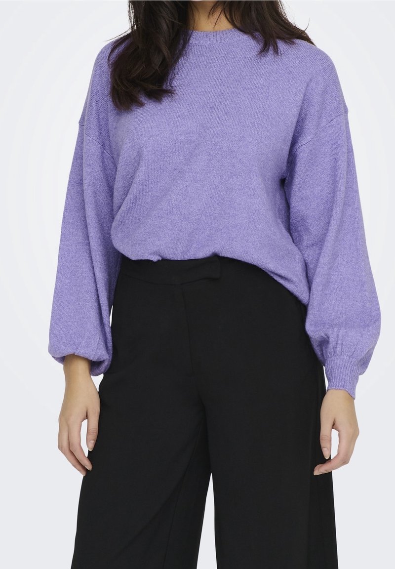 Femme portant un pull lavande ample à manches ballon, rentré dans un pantalon noir taille haute à jambes larges, debout devant un fond blanc.