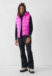 Gilet imbottito di colore rosa acceso con cappuccio e chiusura con zip, indossato sopra una maglia nera a maniche lunghe. Pantaloni neri con righe laterali gialle. Stivali beige massicci.