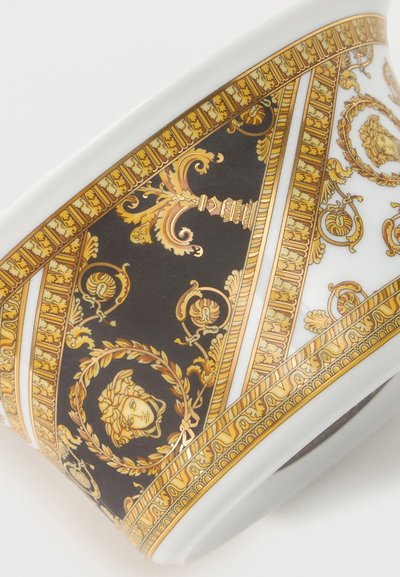 Λευκό κεραμικό φλιτζάνι με περίτεχνα μαύρα και χρυσά μοτίβα μπαρόκ, που περιλαμβάνουν κεφαλές Μέδουσας και περίπλοκα λουλουδένια και σπείρες μοτίβα.