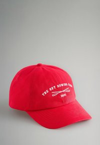 Punane puuvillane nokamüts kummus servaga, millel on valge tikandtekst "THE SET ROWING CLUB" ja kaks ristatud aeru koos "1998" all.