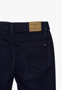 Jeans de mezclilla oscura con un parche de cuero marrón en la cintura, dos bolsillos traseros y un sutil detalle de costura a lo largo de los bordes.