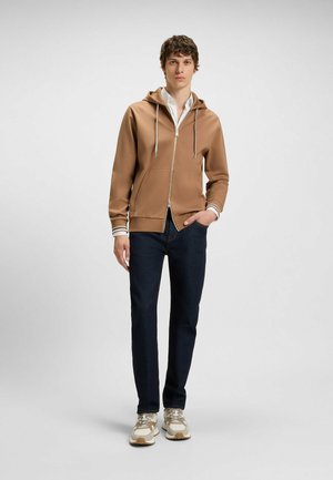 Brown zip-up hoodie with drawstring, front pockets, ribbed white-striped cuffs, paired with dark jeans and beige sneakers.
