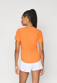 Πορτοκαλί αθλητική μπλούζα με κοντά μανίκια, στρογγυλή λαιμόκοψη, απαλό ύφασμα και μανίκια ράγκλαν, σε συνδυασμό με λευκό σορτς.