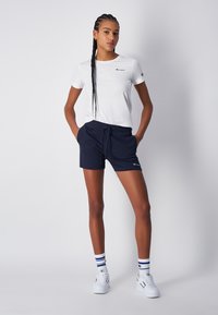 Camiseta blanca de manga corta con un pequeño logo, combinada con pantalones cortos azul marino que tienen una cintura elástica y bolsillos laterales. Zapatillas blancas y calcetines de rayas.