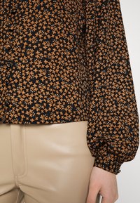 Schwarze Bluse mit kleinem orangen floralem Muster, mit einem lockeren Schnitt und gepufften Ärmeln. Kombiniert mit beigefarbenen, hochgeschnittenen Kunstlederhosen.