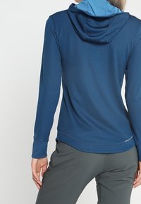 Blå långärmad hoodie i texturerat tyg, med en dragsko kapuschong och en sömlös design. Bakvyn visar släta sömnadsdetaljer.