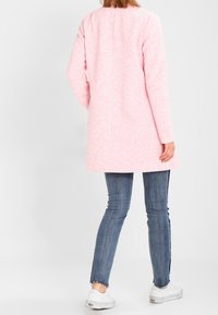 Cappotto lungo texture rosa chiaro con maniche lunghe, scollatura arrotondata e taglio dritto; indossato sopra pantaloni jeans blu aderenti e scarpe bianche.